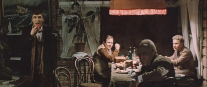 Vintage black-and-white film still shows four men in 1970s attire seated around a wooden table in a dimly lit room with curtains and plants, one man standing nearby smoking a cigarette, bottles and food on the table, red-fringed lamp overhead. Modern color photo depicts a middle-aged man with gray hair smiling while wearing a blue suit and white shirt, standing outdoors against a blurred orange building and green hedges. qqziddxiqreixxkmp